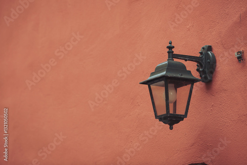 A vintage black lantern is attached to an orange wall, with the soft glow of light illuminating the surroundings in a quiet urban setting during the late afternoon