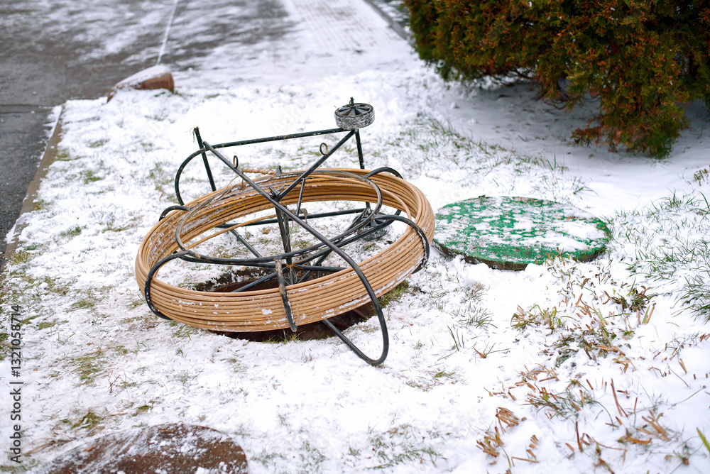Fiber optic cable reel placed near an open maintenance hole during broadband network installation on snowy day in telecommunications engineering site. Coiled cable on metal frame. Selective focus