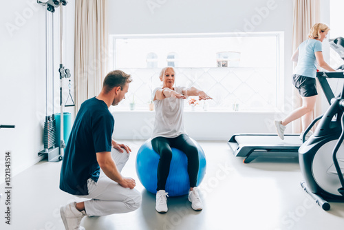 Elderly woman performing balance exercises on a stability ball under supervision of a male trainer in a bright, modern gym setting