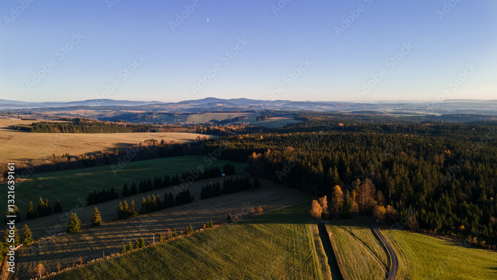 Fototapeta premium Aerial view of a vast countryside at sunset, with rolling hills, dense forests, and open fields. The golden light casts long shadows, highlighting the beauty of the landscape. Orlické hory, Czechia.