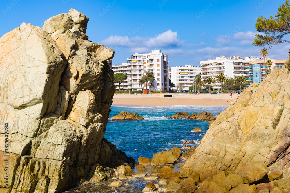 Formaciones rocosas enmarcan la vista de la Playa de Fenals en Costa Brava, con mar azul y edificios.