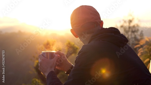 Back view of male hiker wearing a cap, holding a steaming mug and enjoying a peaceful moment at sunrise over a beautiful mountain landscape