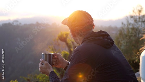 Male hiker wearing orange cap savoring hot beverage at sunrise, overlooking expansive mountain valley with serene landscape and golden morning light