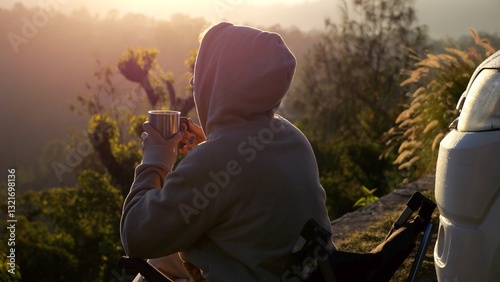 Young camper wearing a hooded sweatshirt sits on a folding chair, sipping from a metal mug and enjoying the sunrise over tranquil mountain valley next to their van. Concept morning with cup of coffee