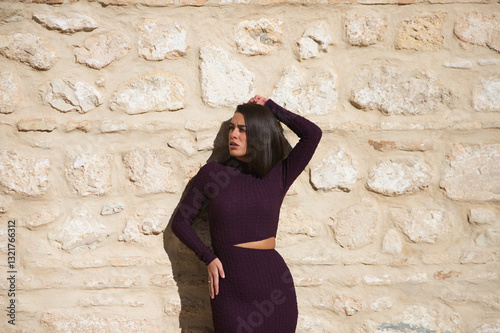 Beautiful young woman dressed in elegant violet dress posing on a stone wall. The woman has the sun on her face and raises one arm above her head and looks to the side. photo