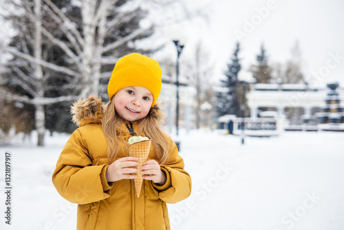 A little girl in a yellow jacket and hat holding an ice cream in her hand and smiling. Winter walk in a city park. 