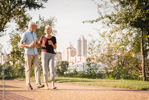 Happy senior couple family walking in summer city park enjoying retirement and spending time together in nature drinking coffee. Romance, love, care and elderly people relationship concept, copy space