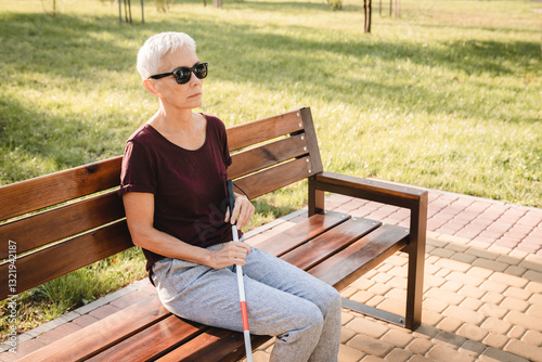 Konstfotografi An elderly caucasian visually impaired woman in black casual sunglasses sitting wooden bench in park with white folded tactile cane stick in hands
