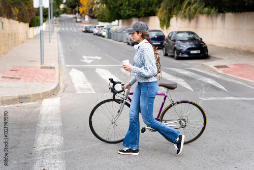 Young woman walking with bicycle and coffee across crosswalk