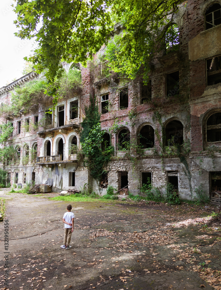 Abkhazia. Child kid stands against old building overgrown by green trees and grass in summer. Concept of war