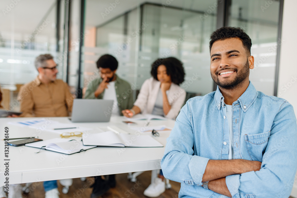 Fototapeta premium A young businessman in a casual denim shirt sits confidently at a modern workspace, exuding positivity while colleagues engage in a team discussion.