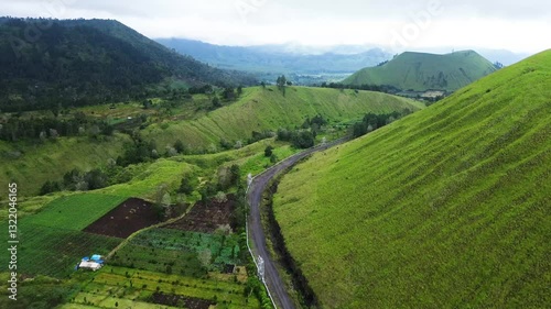Stunning aerial footage tracking winding roads through vast green hills and geothermal power pipelines, surrounded by misty grasslands in Wurung Crater, Indonesia.