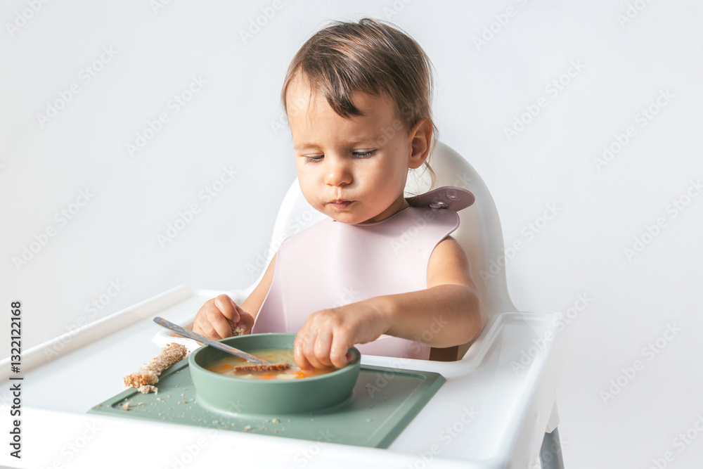 Toddler sitting in a high chair and dipping a piece of bread into a bowl of soup, wearing a silicone bib, focused on eating in a bright and minimalistic home setting Toddler sitting in a high chair and dipping a piece of bread into a bowl of soup, wearing a silicone bib, focused on eating in a bright and minimalistic home setting