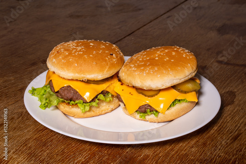 Two cheeseburgers with beef patties, melted cheese, lettuce, and pickles on sesame buns, served on a white plate atop a rustic wooden surface.