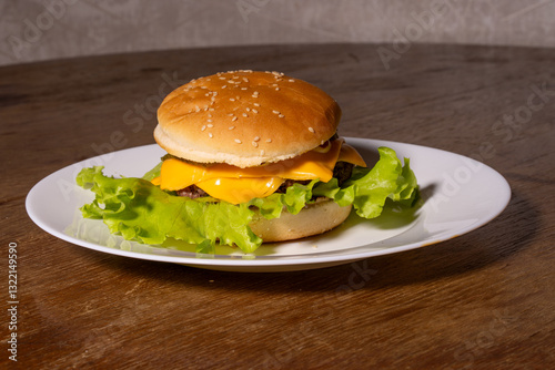 Obraz na płótnie A juicy cheeseburger with a sesame seed bun, melted cheese, beef patty, and lettuce on a white plate, styled atop a rustic wooden background. w salonie
