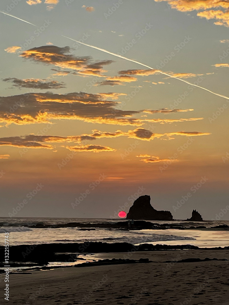 sunset over the sea with a red sun by a shark tooth looking rock with clouds and a trail left behind from a plane