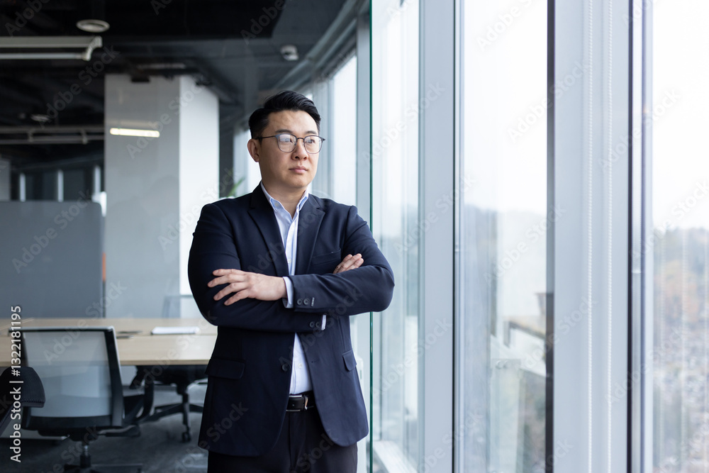 Asian young male businessman standing in the office near the window wearing a suit and glasses, arms crossed on his chest and looking to the side Asian young male businessman standing in the office near the window wearing a suit and glasses, arms crossed on his chest and looking to the side