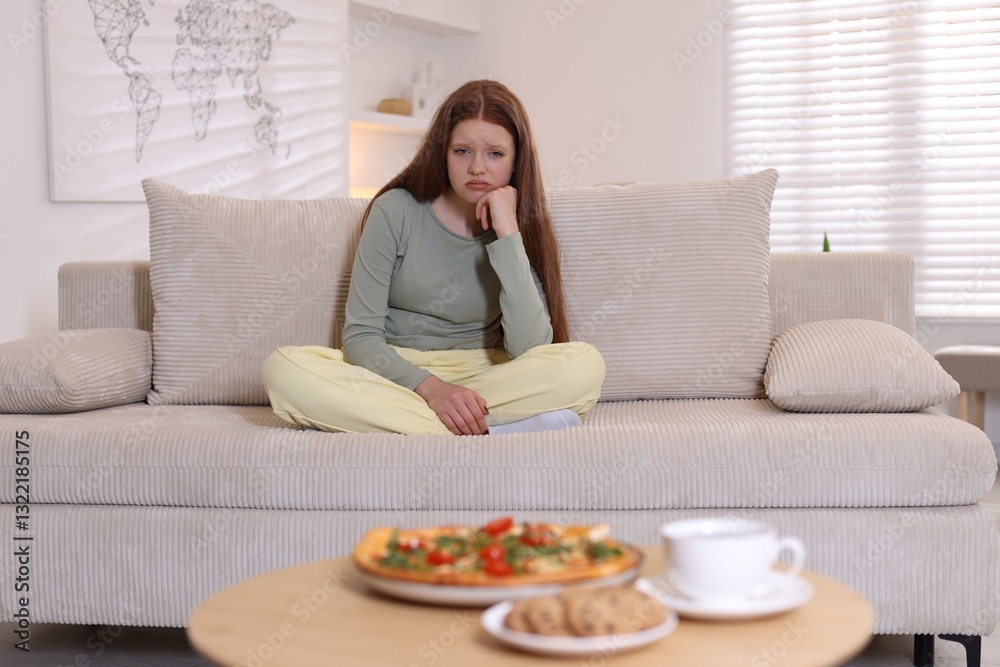 Sad teenage girl sitting on sofa near table with pizza, coffee and cookies at home. Eating disorder Sad teenage girl sitting on sofa near table with pizza, coffee and cookies at home. Eating disorder