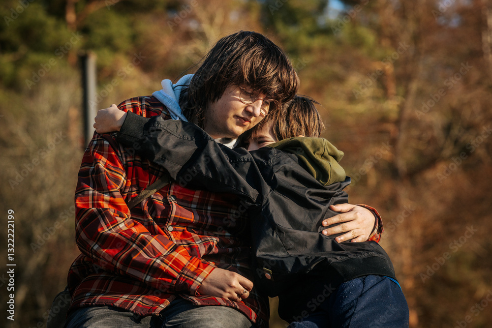 A young person in a plaid jacket and glasses embraces a child in a black jacket and green hoodie. The warm sunlight highlights their emotional connection in an outdoor setting. A young person in a plaid jacket and glasses embraces a child in a black jacket and green hoodie. The warm sunlight highlights their emotional connection in an outdoor setting.