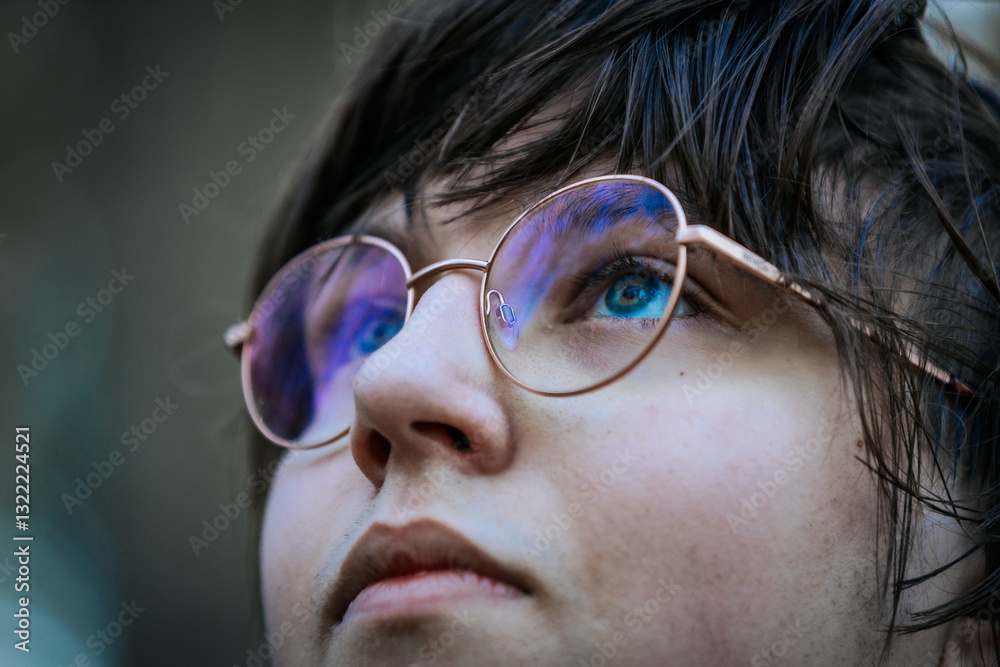 A close-up of a person wearing round glasses with a gold frame, reflecting light. Their blue-green eyes gaze upwards, creating a thoughtful and introspective atmosphere. A close-up of a person wearing round glasses with a gold frame, reflecting light. Their blue-green eyes gaze upwards, creating a thoughtful and introspective atmosphere.