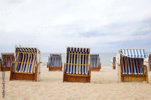 Fototapeta Naklejka Na Ścianę i Meble -  Wicker beach baskets with white-blue striped fabric rest on Kolobrzeg’s sandy shore under a calm Baltic sky.