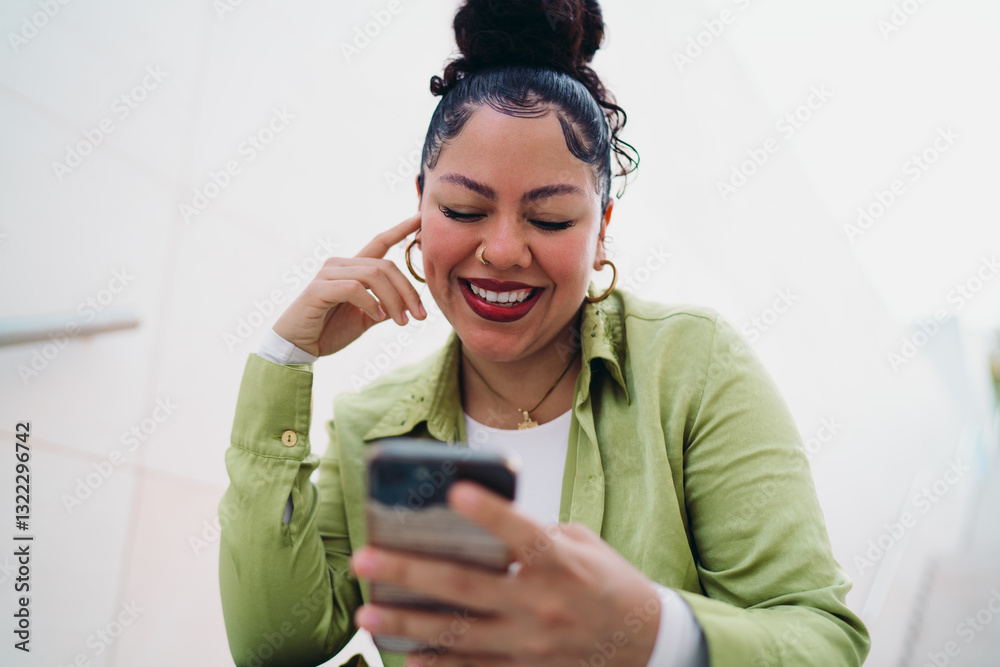 Fototapeta premium Young woman in green shirt sits outdoors, smiling while looking at her smartphone app. Female appears amused, likely watching funny videos or engaging on social media. Digital entertainment concept