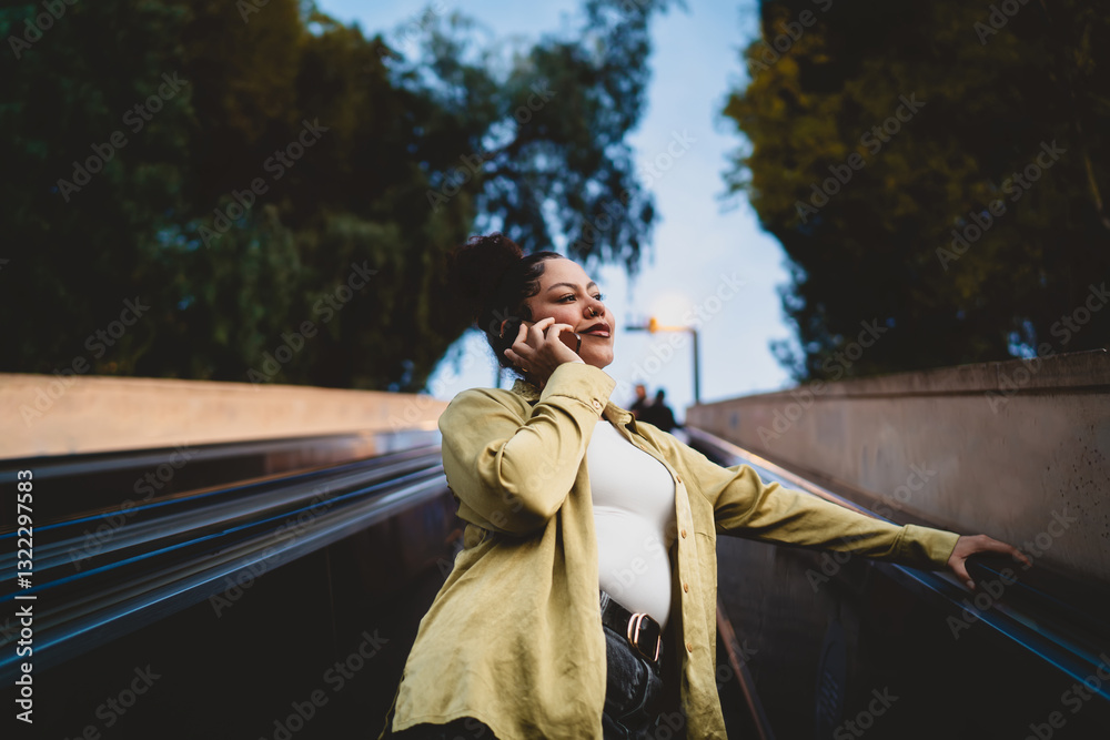 Obraz premium young Hispanic woman smiles while talking on phone as she rides an outdoor escalator at dusk. Dressed in green shirt, female looks content and engaged in conversation, with city lights in background