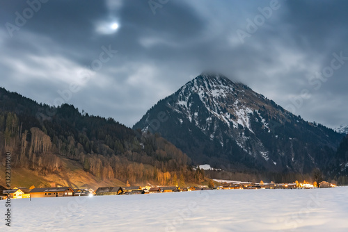 Obraz na płótnie Winter night landscape in the mountains, full moon hidden behind clouds over the mountains. Small village in the Alps, houses lit at night, view from a hiking trail.at night, view from a hiking trail. w salonie
