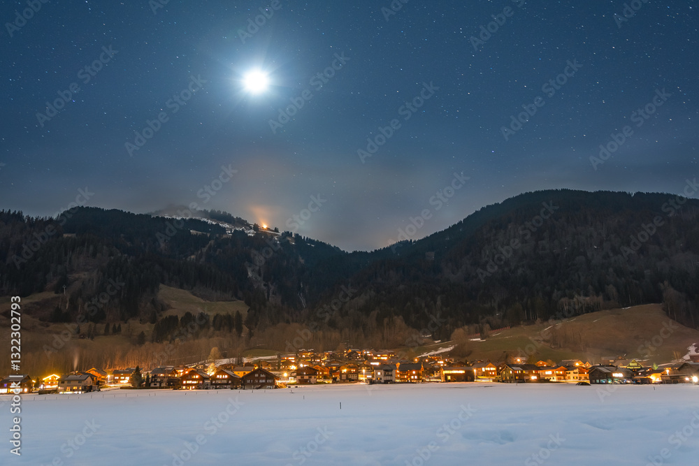 Winter night landscape in the mountains, full moon in the starry sky above the mountains. Small village in the Alps, houses illuminated at night, view from a hiking trail.