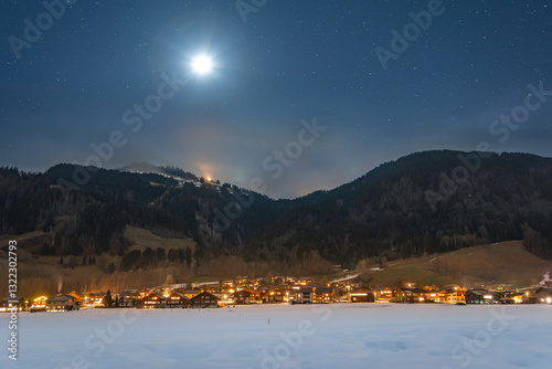 Obraz na płótnie Winter night landscape in the mountains, full moon in the starry sky above the mountains. Small village in the Alps, houses illuminated at night, view from a hiking trail. w salonie