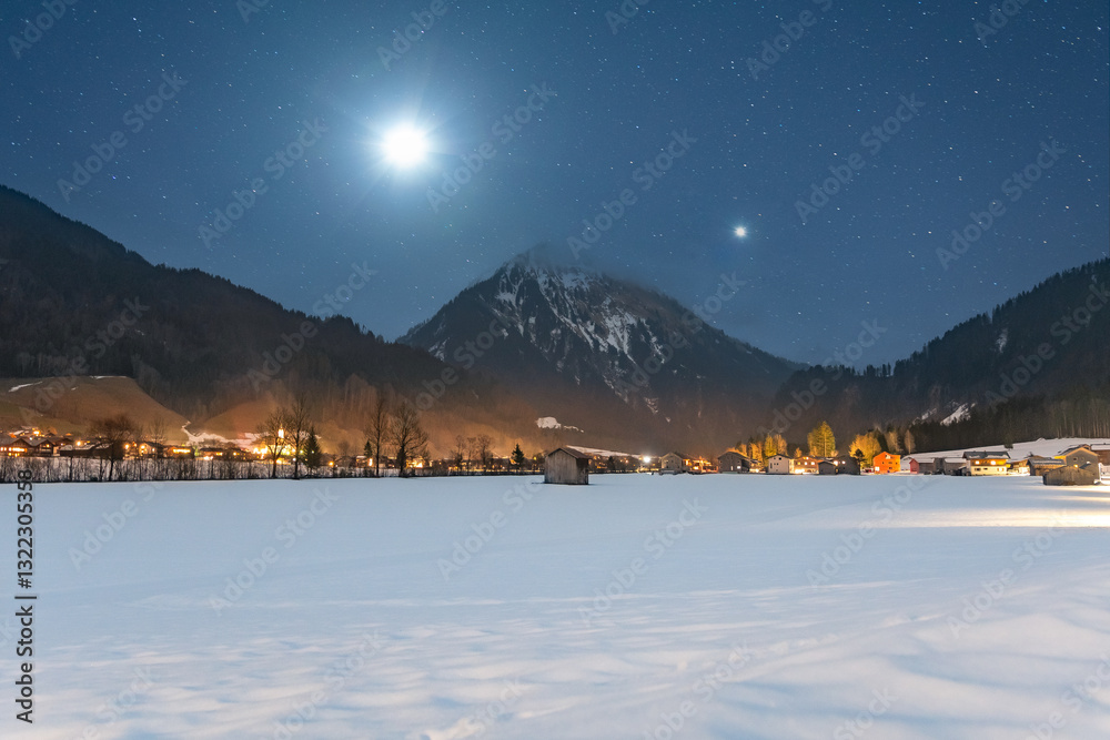 Winter night landscape in the mountains, full moon and planet Venus in the starry sky above the mountains. Small village in the Alps, houses illuminated at night, view from a hiking trail.