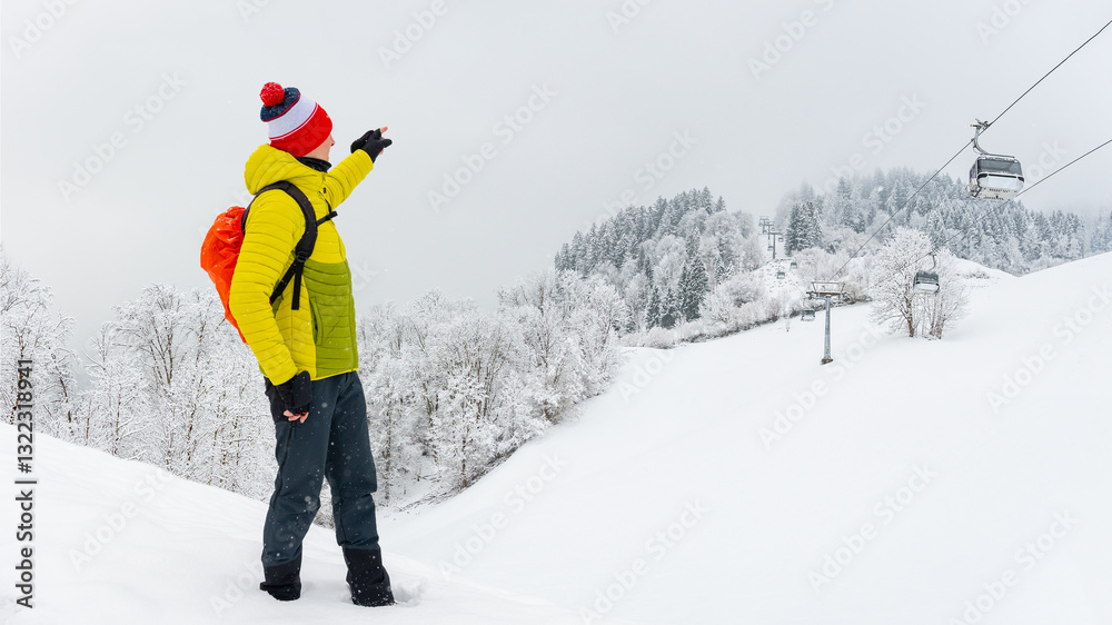 A young man stands on the trail and shows how high the gondola lift goes. Winter landscape on a mountain hiking trail on a cloudy and foggy day.