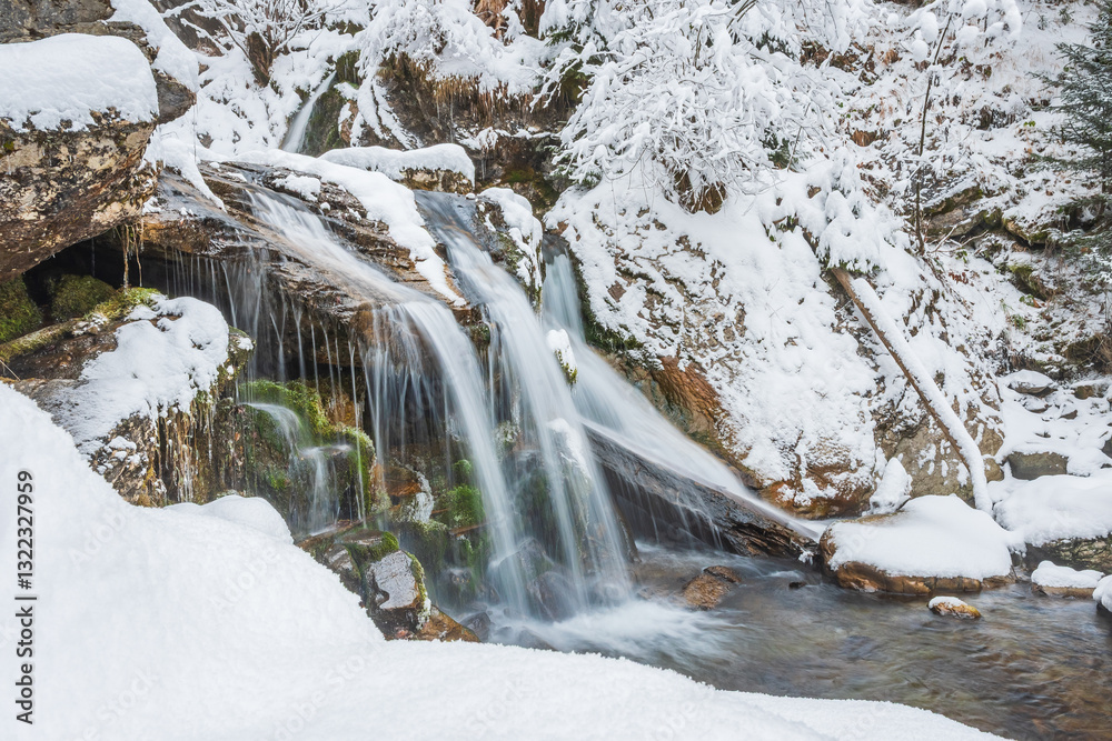 Small waterfall on a mountain river, water flows down snow-covered rocks. Winter landscape on a mountain hiking trail.