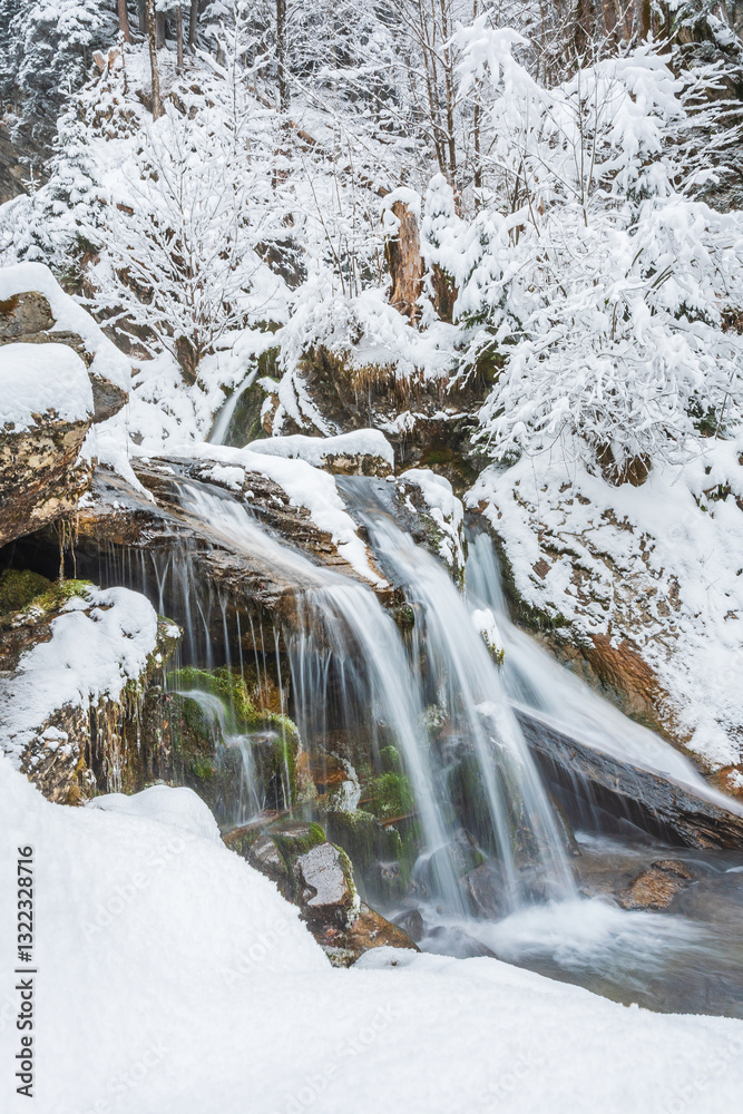 Small waterfall on a mountain river, water flows down snow-covered rocks. Winter landscape on a mountain hiking trail.