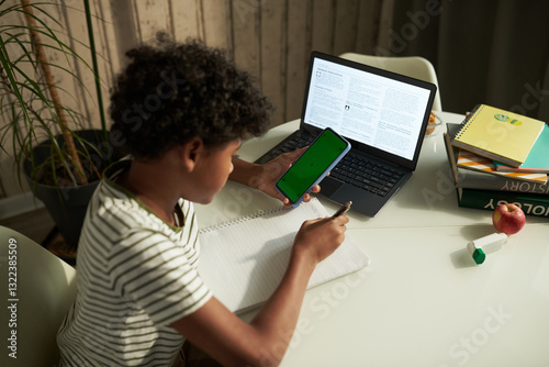 High angle of diligent child taking notes in copybook and using smartphone with chroma key screen in front of laptop