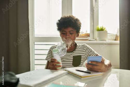 Asthmatic schoolboy with inhaler breathing vapors from mask while sitting by table at home and carrying out schoolwork