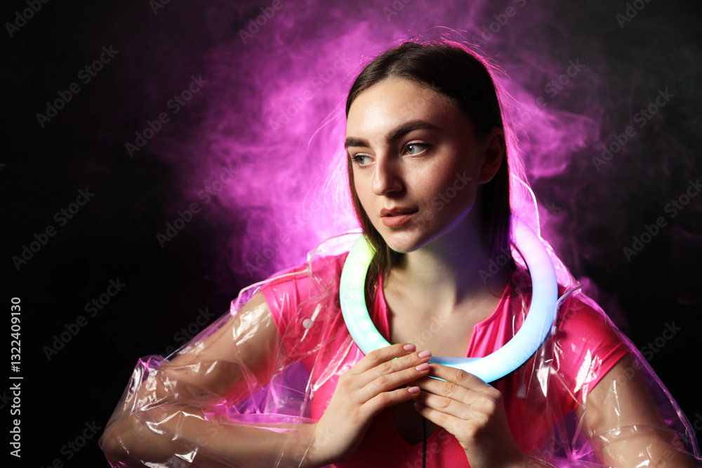Naklejka premium Beautiful woman with ring lamp wearing transparent coat on dark purple background in smoke