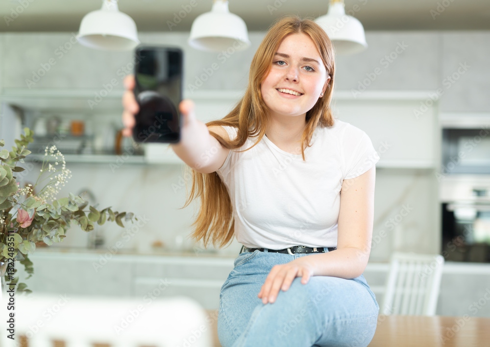 in cozy home interior, smiling woman sits on table and and shows interesting photo on screen of mobile phone