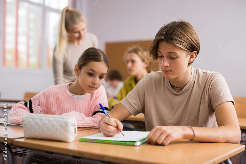 Teenager boy and younger girl sitting at desk in classroom and doing tasks. Teenager boy and younger girl sitting at desk in classroom and doing tasks.