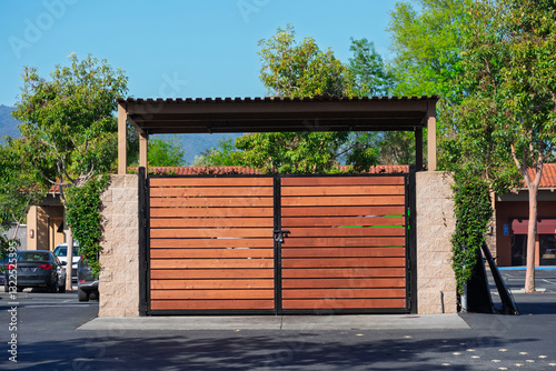 Garbage enclosure with modern wooden slats, stone pillars, and metal roofing, positioned in an urban parking lot with landscaped trees