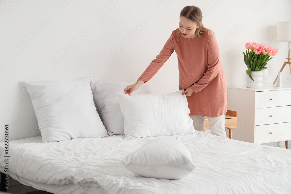 Pretty young woman putting pillow on bed in light bedroom Pretty young woman putting pillow on bed in light bedroom