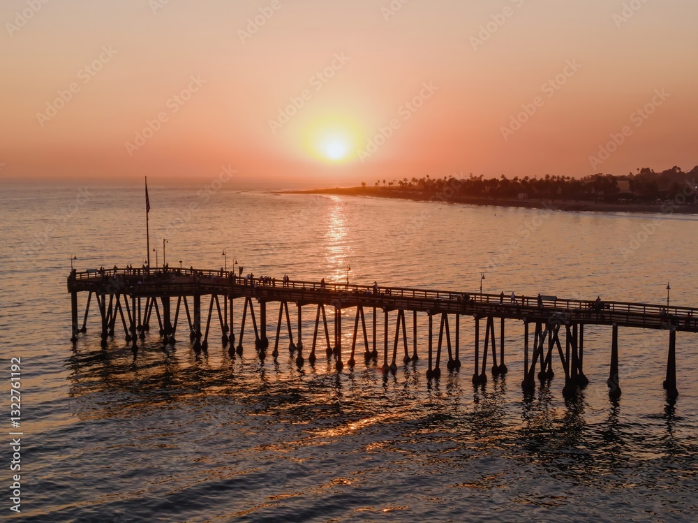 Fototapeta premium People stroll along the pier in Ventura, California, USA, enjoying the sunset. The sun reflects on the water, creating a peaceful and scenic view.
