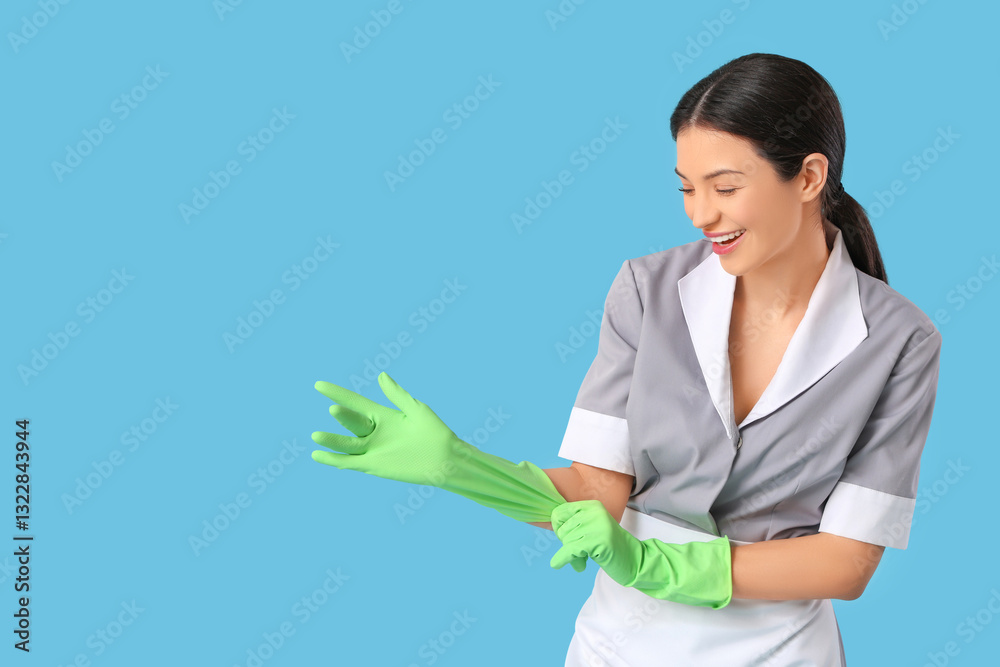 Beautiful young chambermaid putting on rubber gloves on blue background Beautiful young chambermaid putting on rubber gloves on blue background