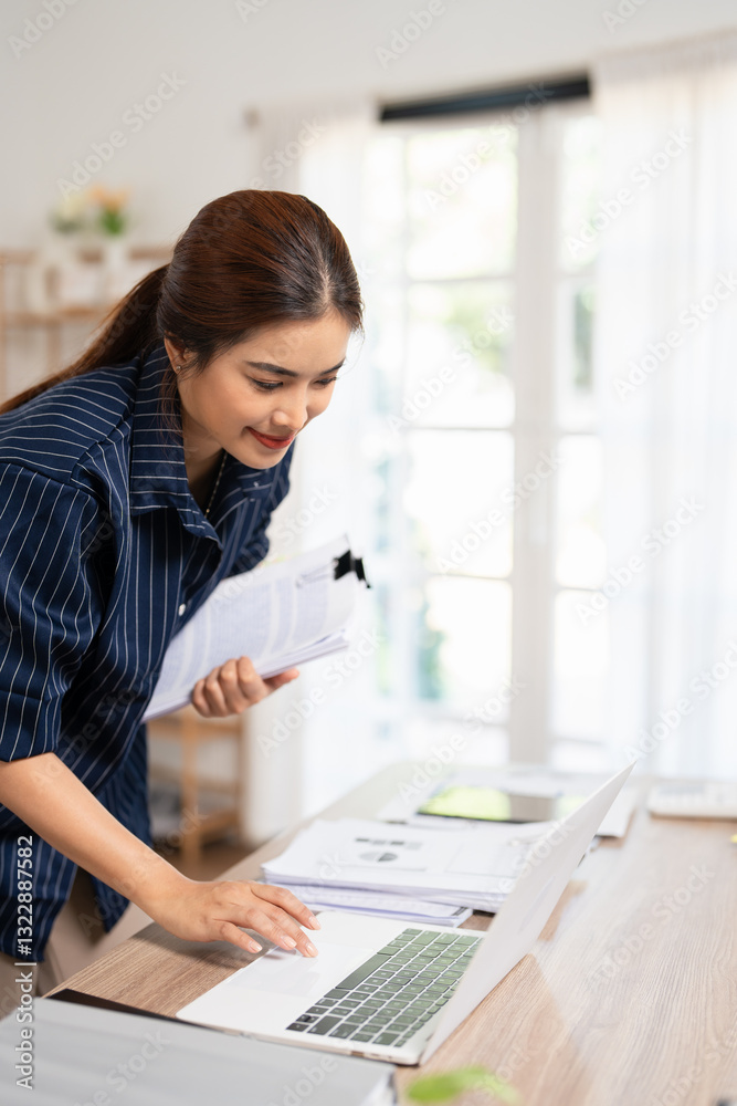 Asian business woman with a stack of papers standing at a desk home office with laptop. focused on finding information . Asian business woman with a stack of papers standing at a desk home office with laptop. focused on finding information .