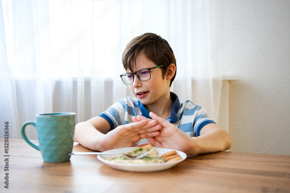 Stubborn boy with a displeased face refuses to eat on a plate, sits in the kitchen without appetite. Upset teenage boy does not want to eat tasteless food, loss of appetite. Stubborn boy with a displeased face refuses to eat on a plate, sits in the kitchen without appetite. Upset teenage boy does not want to eat tasteless food, loss of appetite.