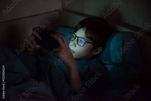 Teenage boy with glasses at night under a blanket with a smartphone. Children who are addicted to gadgets. Mental health problems caused by gadgets. Visual impairment.