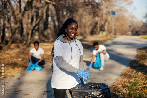 Smiling african american volunteer putting plastic waste into garbage bin with her team of activists.