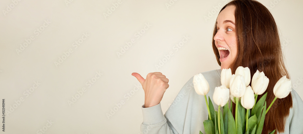 A woman with long brown hair smiles enthusiastically while holding a bouquet of white tulips. She points her thumb to the side, conveying excitement in a light-colored room A woman with long brown hair smiles enthusiastically while holding a bouquet of white tulips. She points her thumb to the side, conveying excitement in a light-colored room