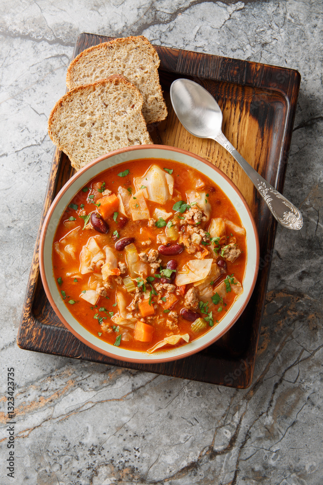 Beef and Cabbage soup is filled with seasoned ground beef, cabbage, carrots, celery, kidney beans, and diced tomatoes closeup in the plate on the table. Vertical top view from above