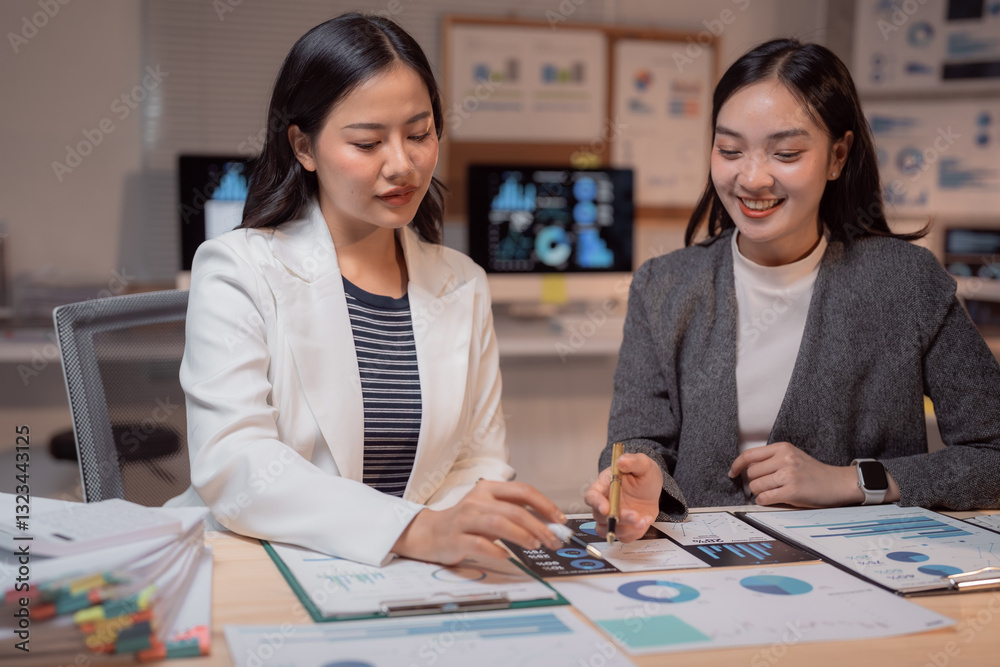 Fototapeta premium Two young Asian businesswomen analyzing financial charts and graphs, working together on a project in a modern office, demonstrating teamwork and dedication